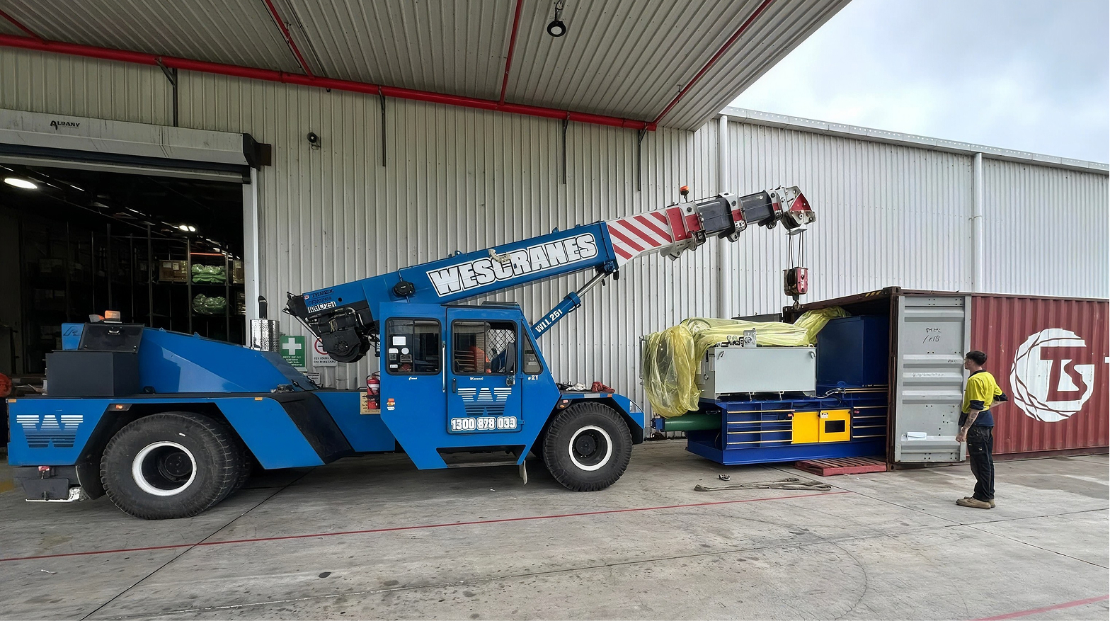 aWescranes crane lowering an industrial baling machine into a shipping container at a warehouse
