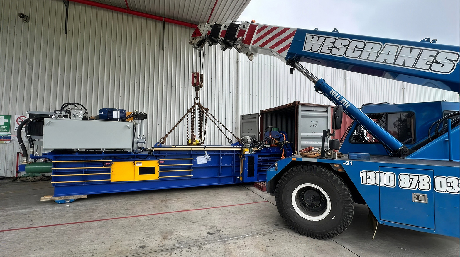 aWescranes crane lifting a large baling machine using chain slings near a shipping container